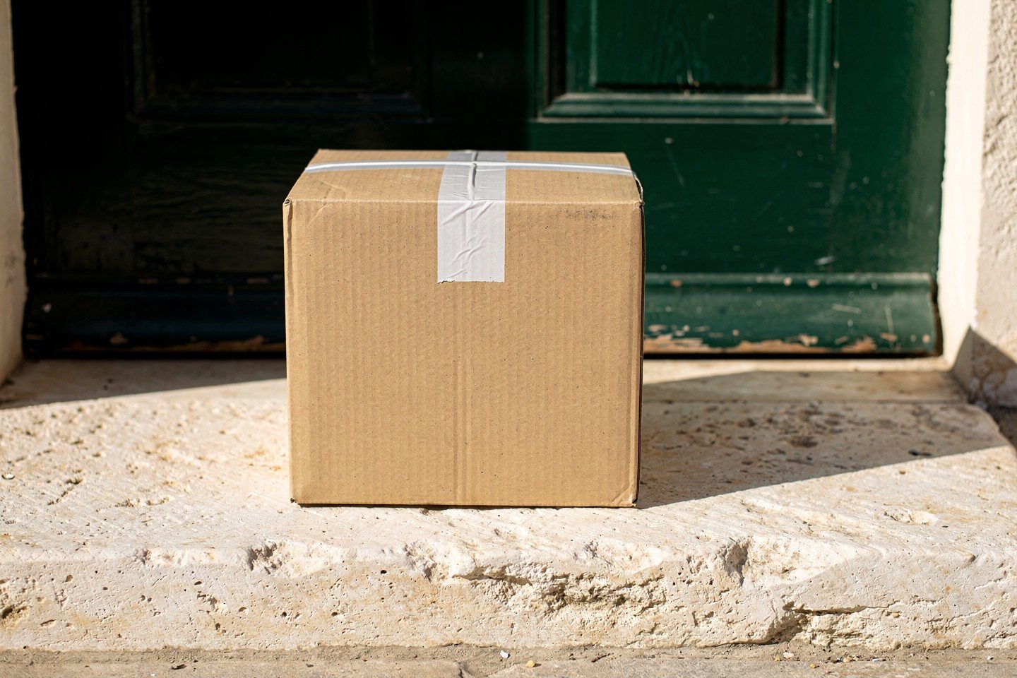 Illustration: a plain unmarked corrugated shipping box on a limestone doorstep in front of a dark green door.